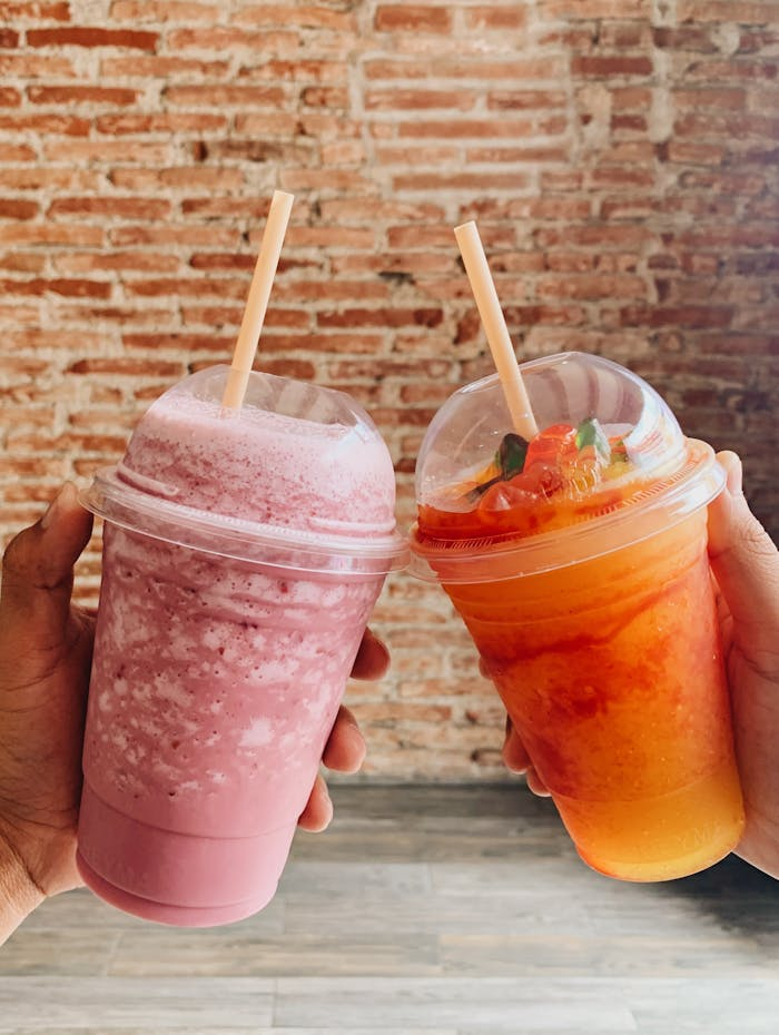 Two colorful fruit smoothies held in hands against a rustic brick wall backdrop.