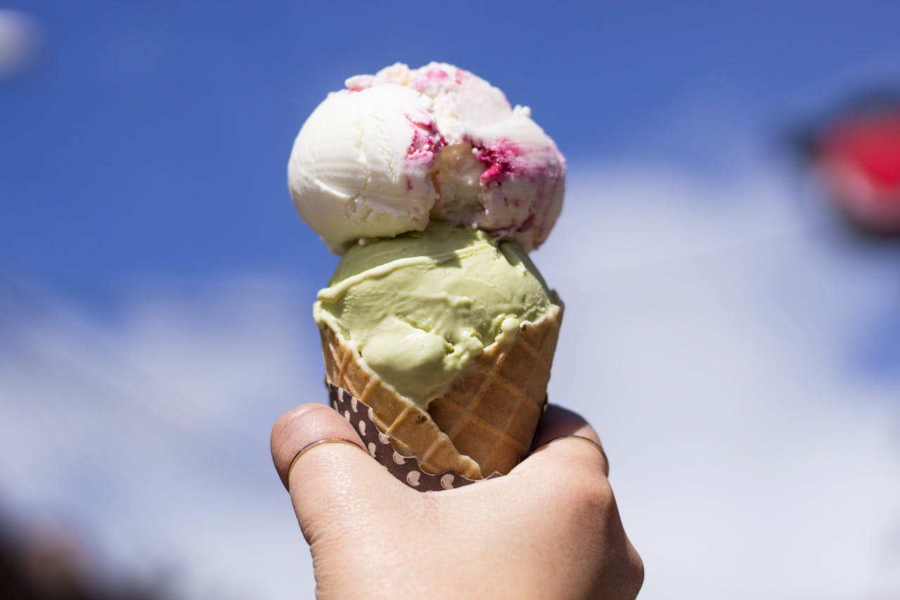 Hand holding a delicious double scoop ice cream cone against a bright blue sky.